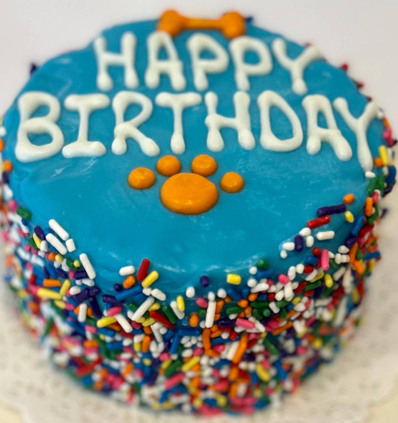 A blue doggie birthday cake with rainbow sprinkles around the side and "happy birthday" written in white on top with a orange bone and paw print.