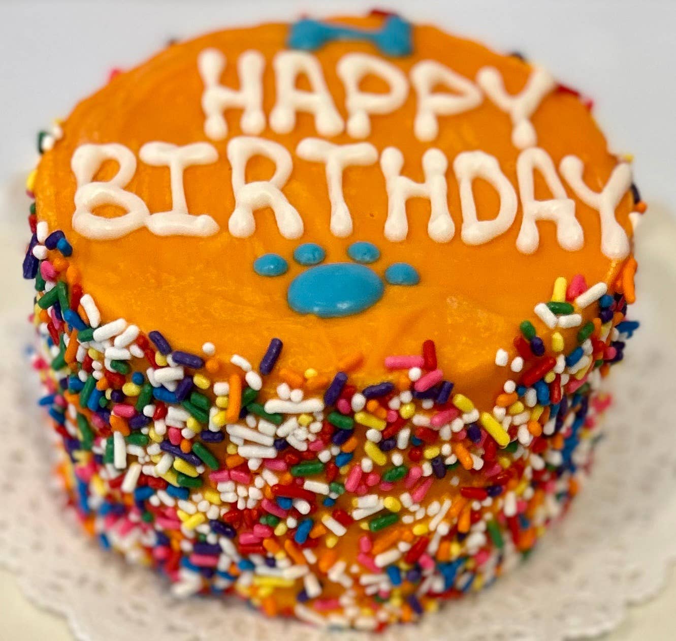An orange doggie birthday cake with rainbow sprinkles around the side and "happy birthday" written in white on top with a blue bone and paw print.