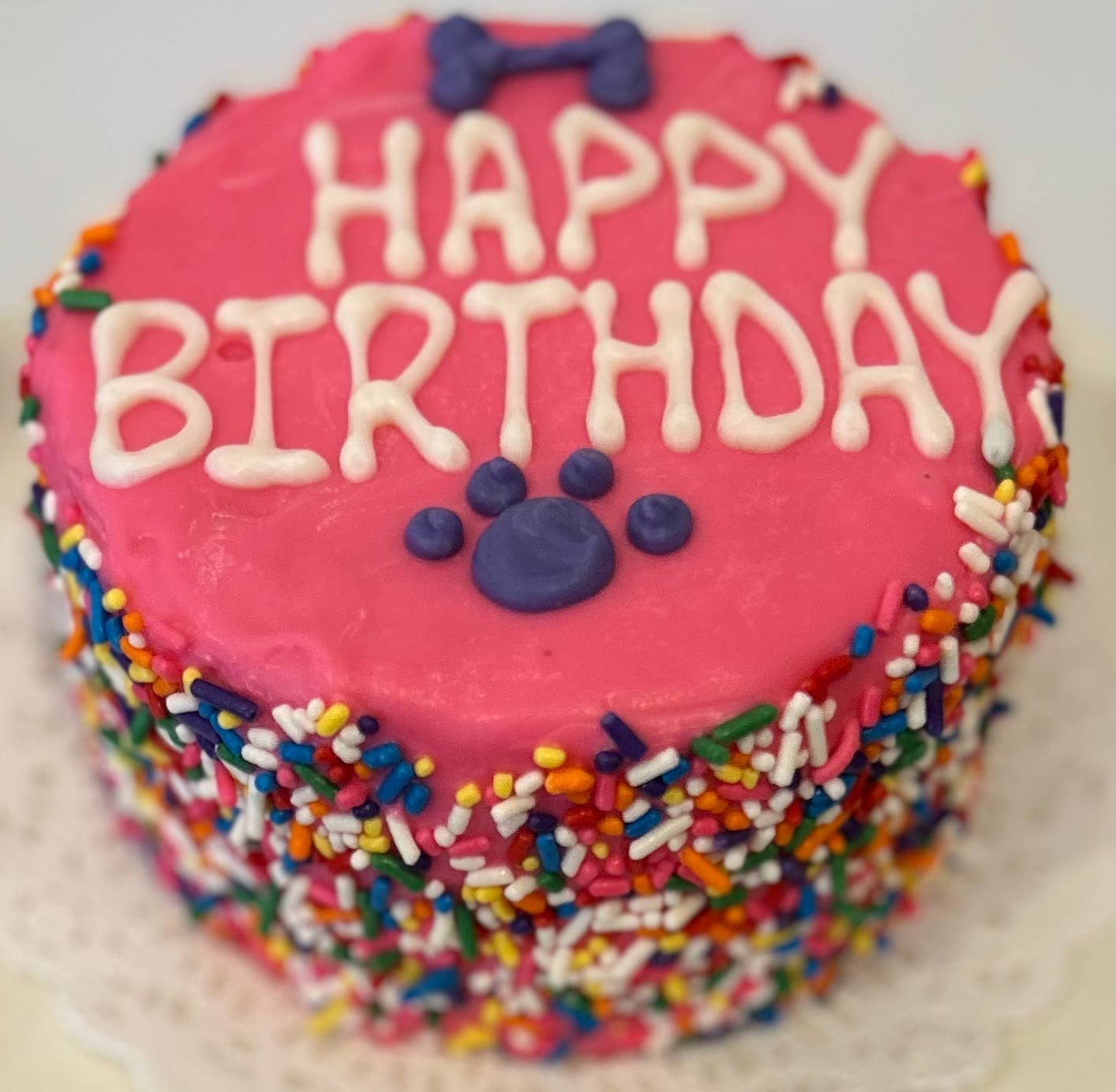 A pink doggie birthday cake with rainbow sprinkles around the side and "happy birthday" written in white on top with a purple bone and paw print.