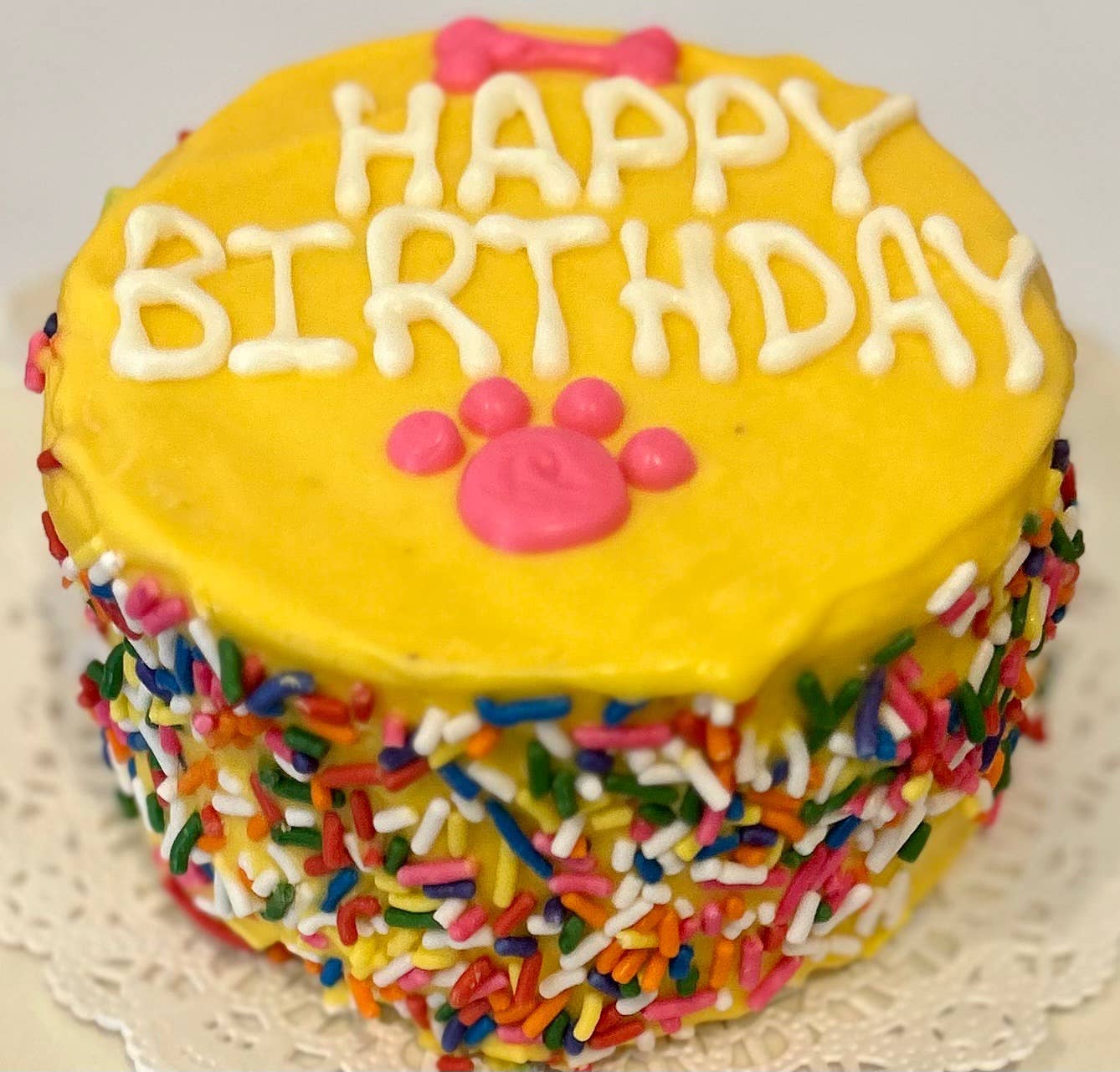 A yellow doggie birthday cake with rainbow sprinkles around the side and "happy birthday" written in white on top with a pink bone and paw print.