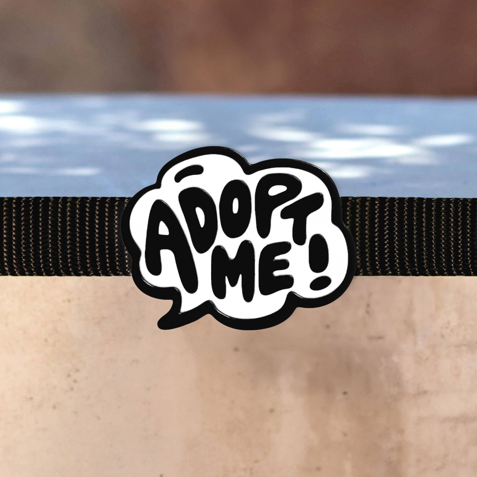 A black and white silicon dog collar charm. It is a cartoon speech bubble that reads, "Adopt Me!"