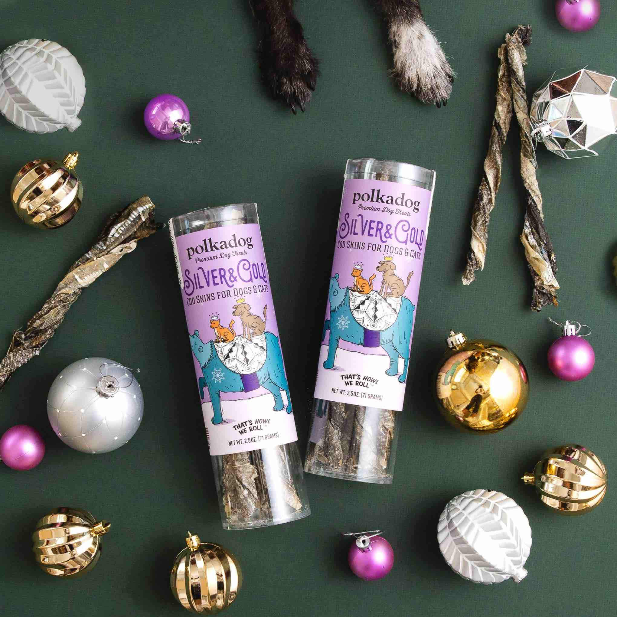 Two tubes of Polkadog cod skin dog treats surrounded by Christmas ornaments on a green background