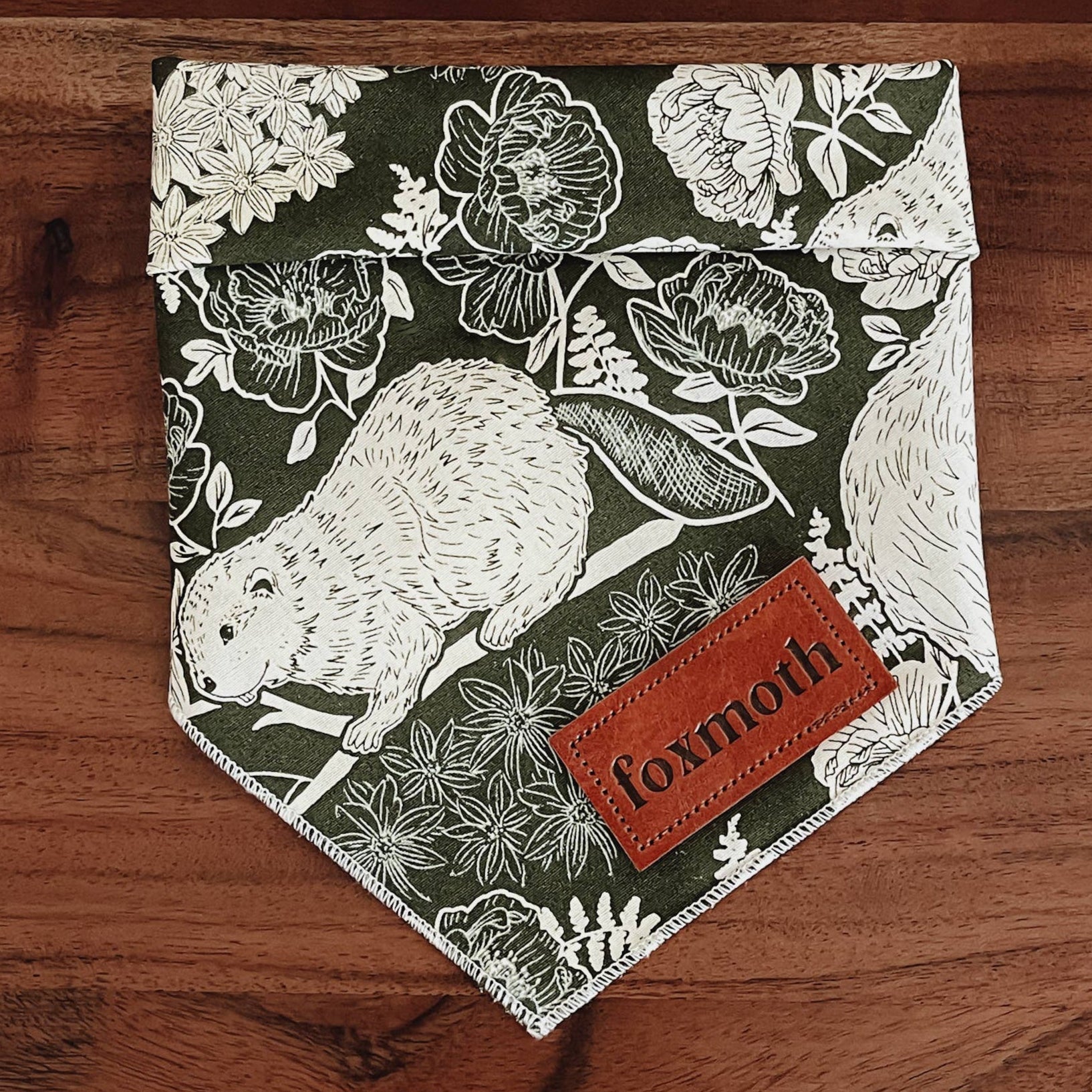 A dog bandana with a floral and beaver print design laid out on a wooden surface.
