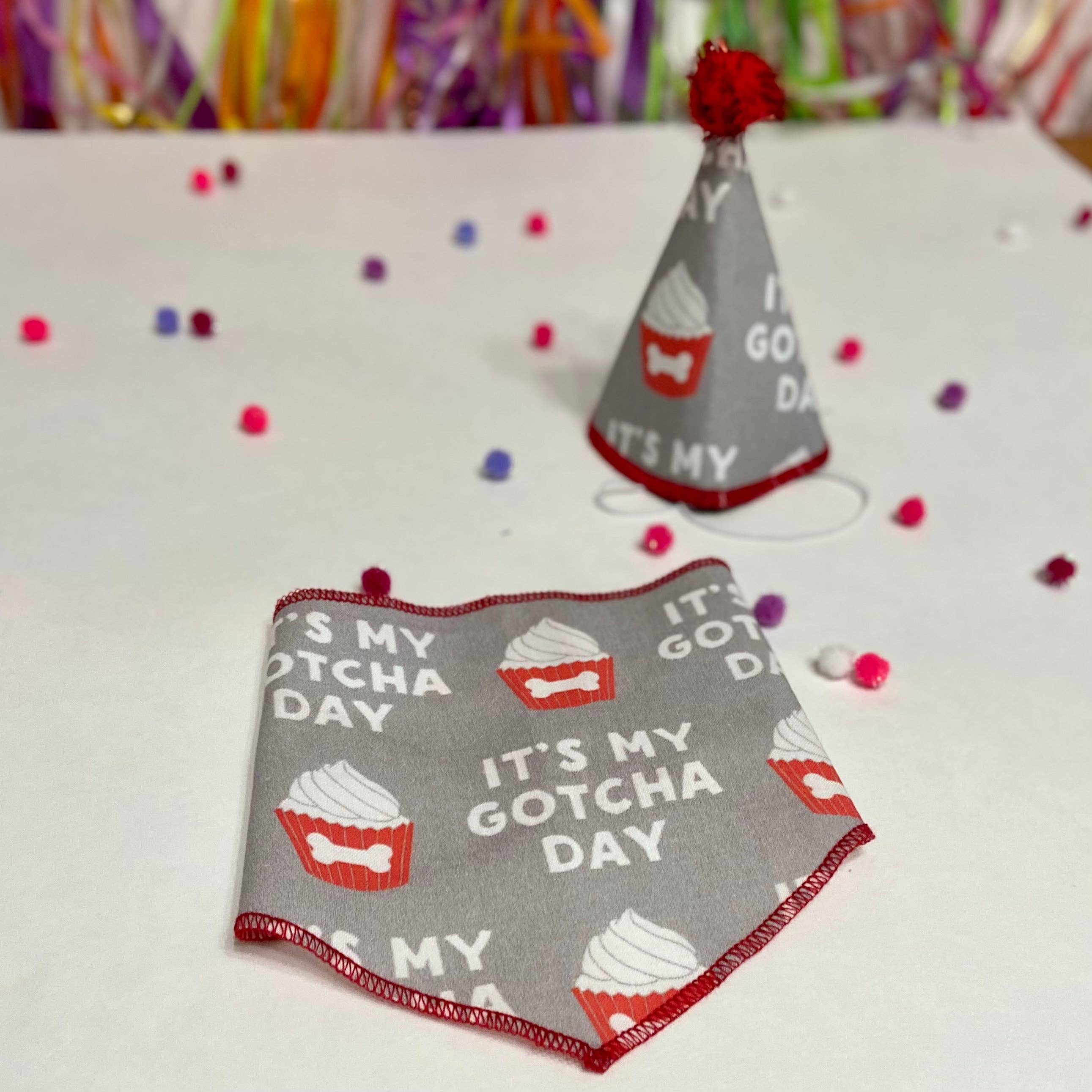Gray pet bib with 'It's My Gotcha Day' text and cupcake design, placed on a white surface with confetti.