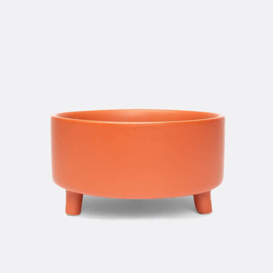 A terracotta, three-legged, elevated, ceramic dog bowl on a white background.