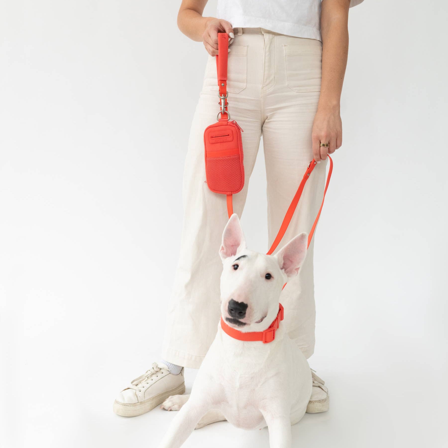 A woman holding a red leash with walk bag attached. On the leash is a white pit bull wearing a matching red collar.