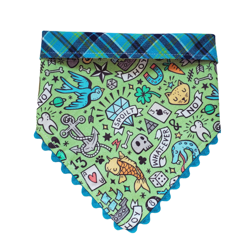 A reversible dog collar bandana with a green and blue plaid pattern on one side and a colorful dog-related tattoo design on the other, with decorative trim around the edges.