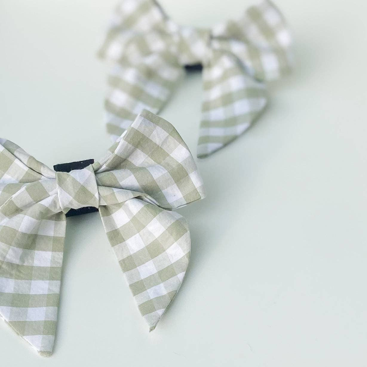 Two sage green gingham dog bows with tails on a white background.
