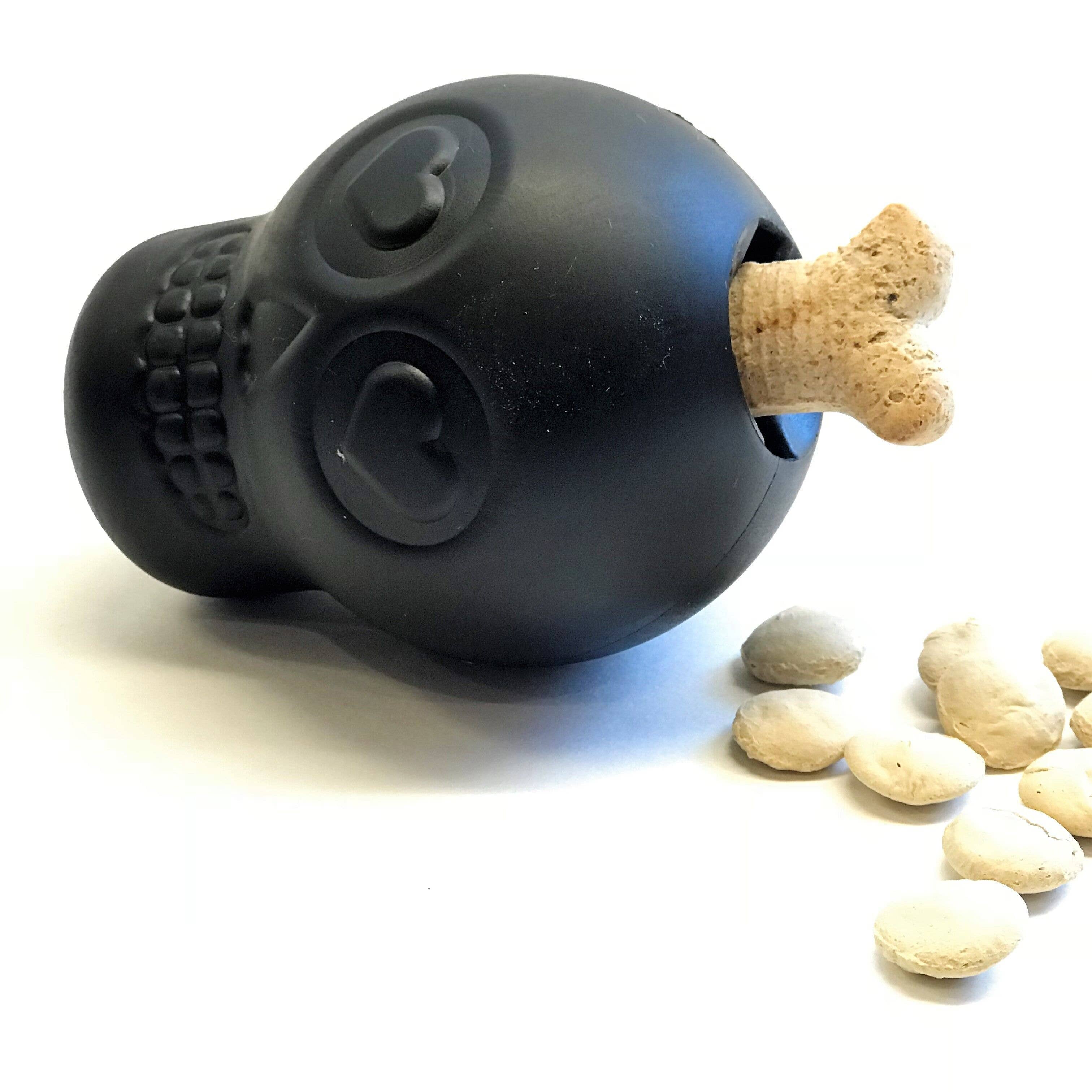 Black, skull-shaped dog chew toy and treat dispenser on a white background with treats.