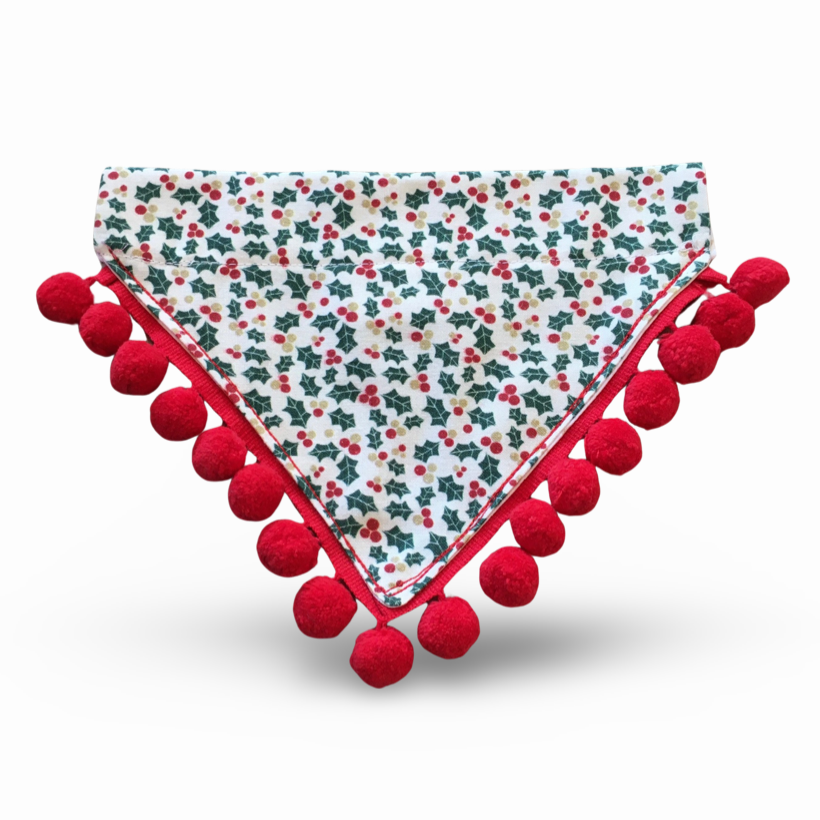 A white bandana with a holly and berries print, trimmed with red pompoms, designed for dogs.