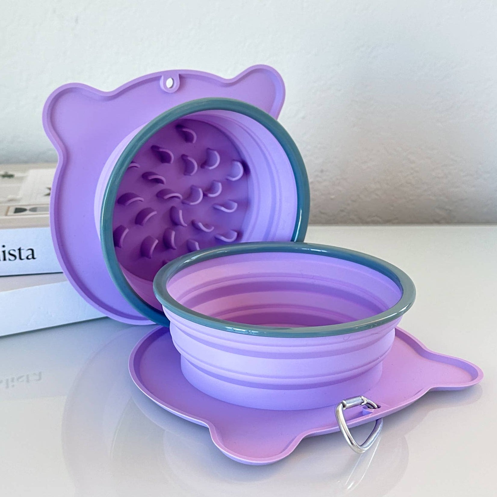Lilac-purple silicone travel bowl with slow feeder feature shown. It's on a white surface with a book in the background.